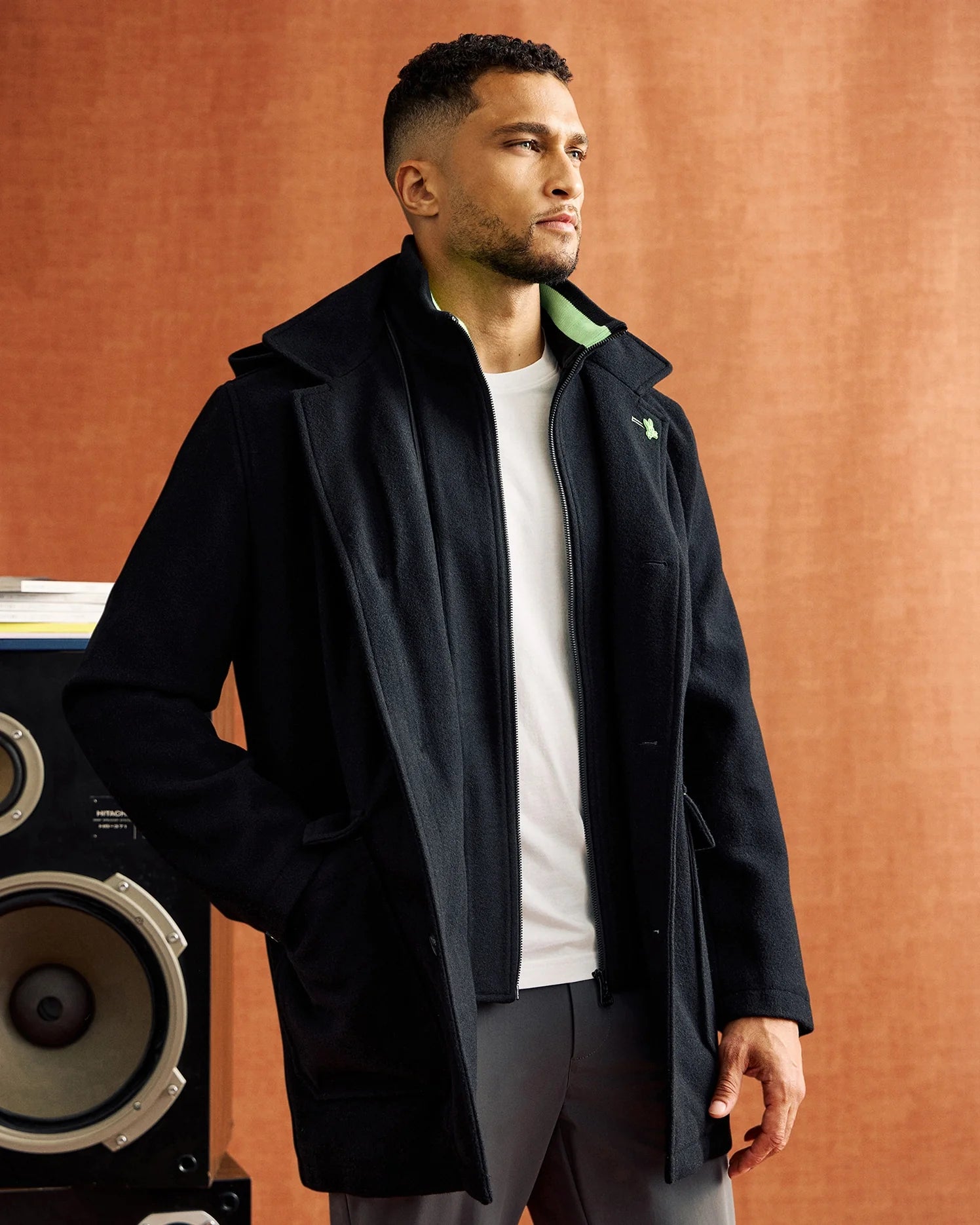 A man stands against a brown backdrop, wrapped in a Psycho Bunny MENS STAFFORD WOOL BLEND COAT - B6N551D200 over a white shirt. He gazes thoughtfully to the side, with a large speaker partially visible behind him.