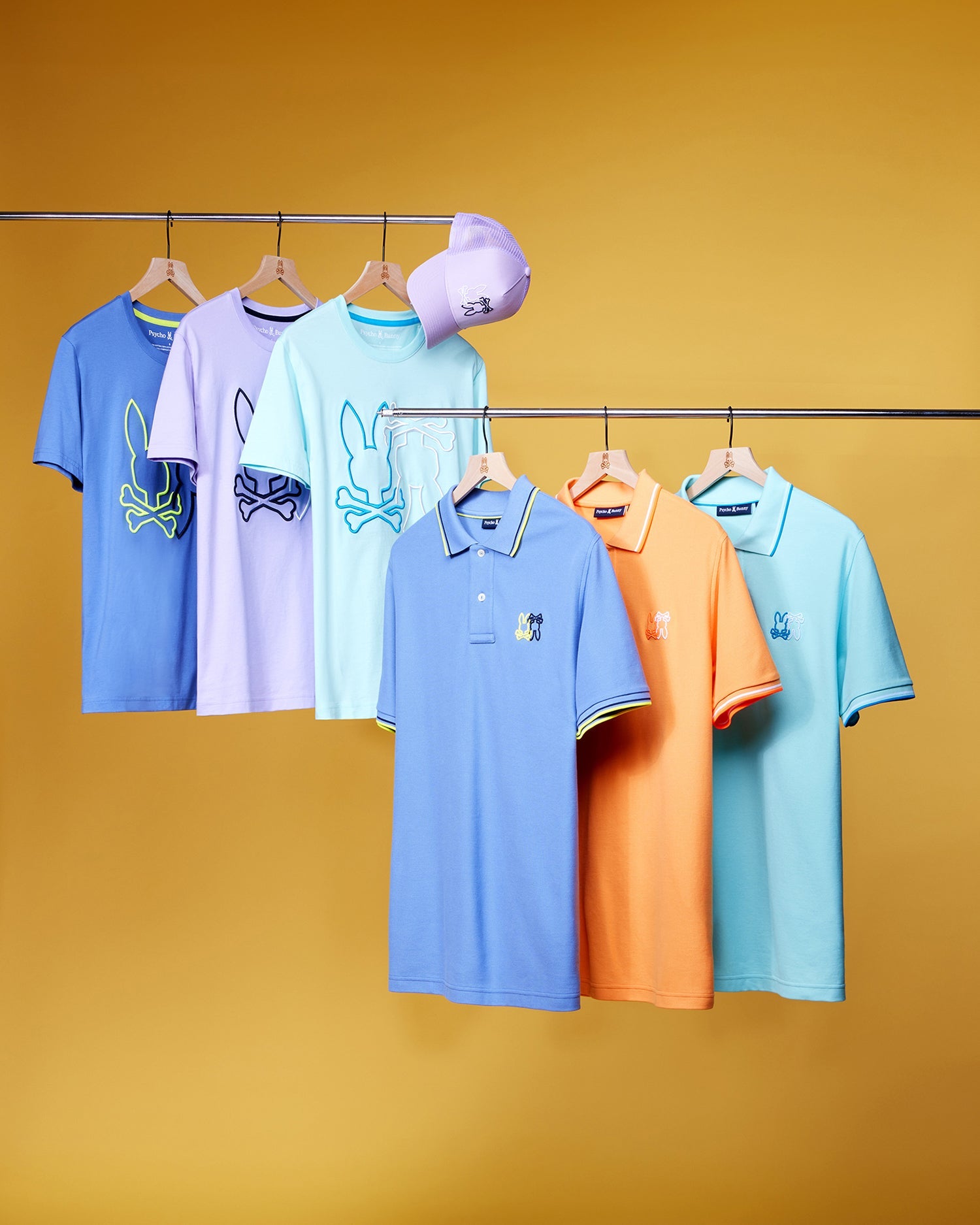 An assortment of five colorful shirts, crafted from Peruvian Pima cotton, hangs from a clothing rack against a yellow background. Among them are three t-shirts and two Psycho Bunny MENS WINTON PIQUE POLO SHIRT - B6K841A2PC. A purple cap rests on the rack. The shirts feature playful designs, including abstract graphics and a rabbit motif.