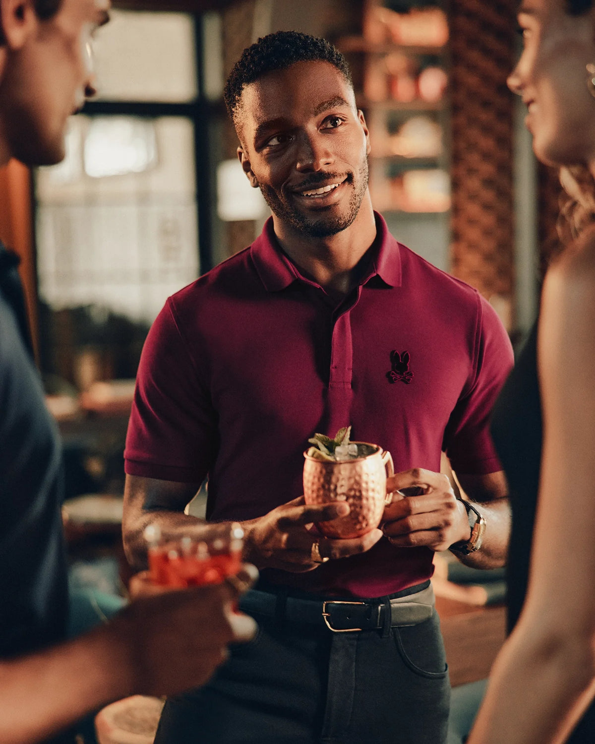 A man wearing a luxurious Psycho Bunny MENS BRANSON PIQUE POLO - B6K108E200 smiles while holding a copper mug with a mint garnish. He engages in conversation with two people, a man and a woman, inside a warmly lit venue.