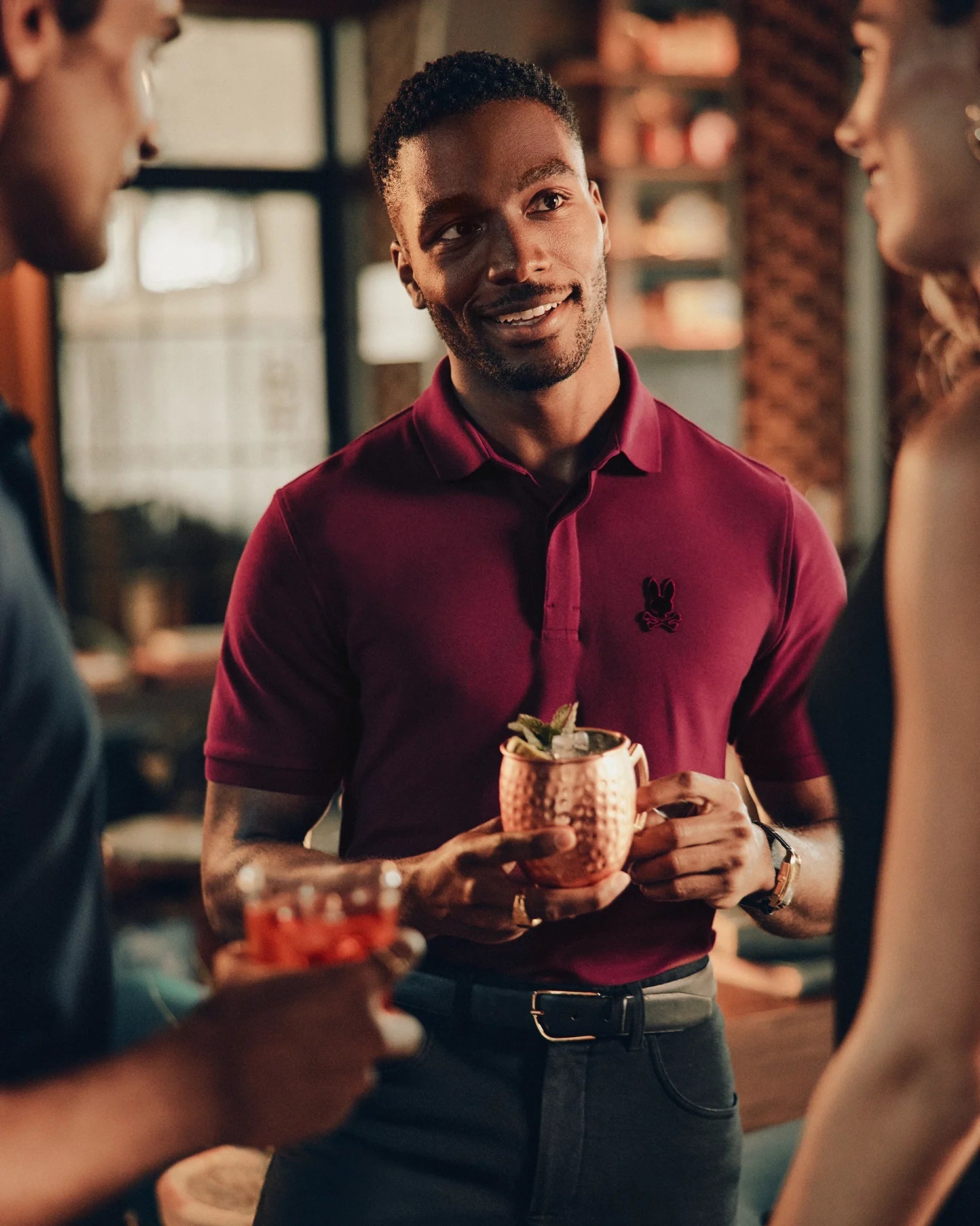 A man wearing a luxurious Psycho Bunny MENS BRANSON PIQUE POLO - B6K108E200 smiles while holding a copper mug with a mint garnish. He engages in conversation with two people, a man and a woman, inside a warmly lit venue.