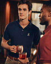 Two men are standing indoors, engaged in conversation. Alexander, holding a copper mug and wearing the Psycho Bunny Men's Alexander Pique Polo (B6K101E200) in navy, chats with the other man, who is partly visible and holds a glass with a red drink. The setting has warm lighting and exudes a relaxed atmosphere.
