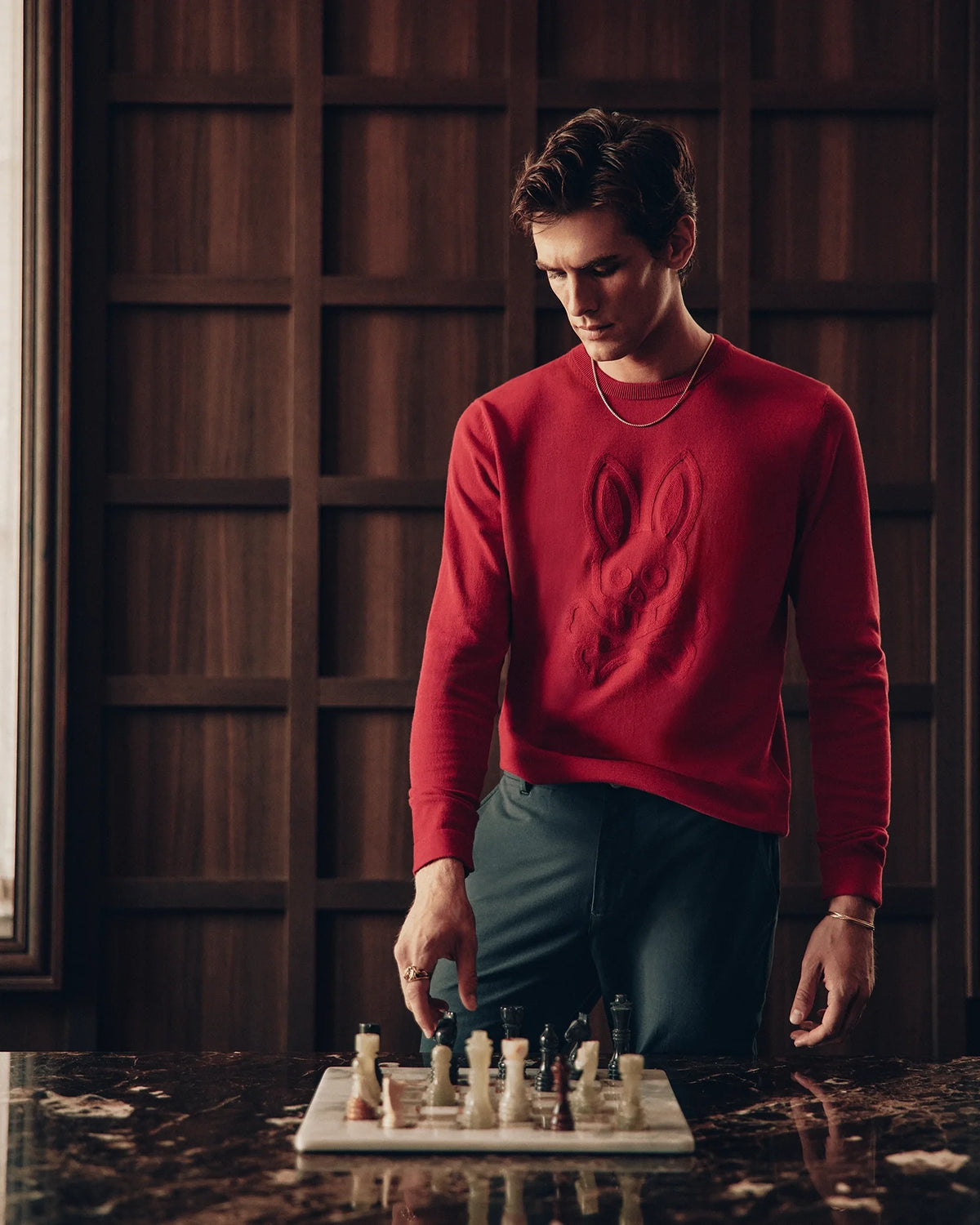 A person in a red Psycho Bunny MENS COORY CHAINSTITCH COTTON SWEATER stands by a marble table with a chessboard, thoughtfully placing their hand near the pieces. The backdrop showcases a wooden wall adorned with geometric patterns.