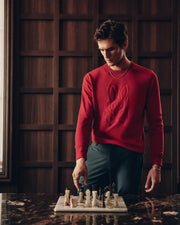A person in a red Psycho Bunny MENS COORY CHAINSTITCH COTTON SWEATER stands by a marble table with a chessboard, thoughtfully placing their hand near the pieces. The backdrop showcases a wooden wall adorned with geometric patterns.