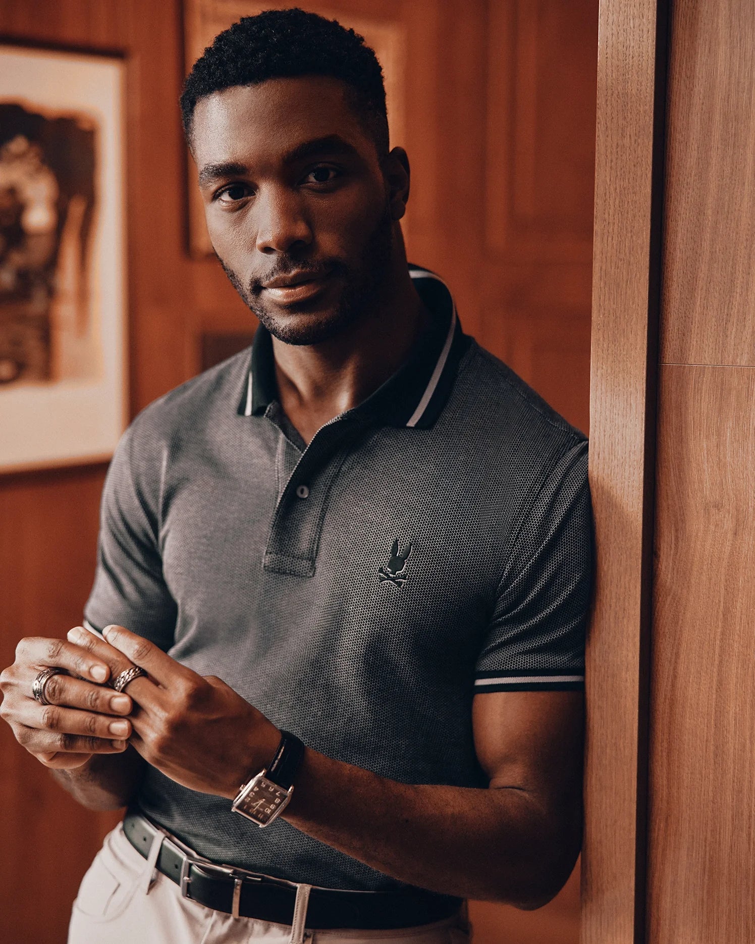A man with short hair and a beard is leaning against a wooden wall, dressed in a dark Psycho Bunny MENS WARSAW JACQUARD PIQUE POLO - B6K901E200 made from Peruvian Pima cotton, light pants, and a watch. He has rings on his fingers and gazes directly at the camera, with framed pictures in the background.